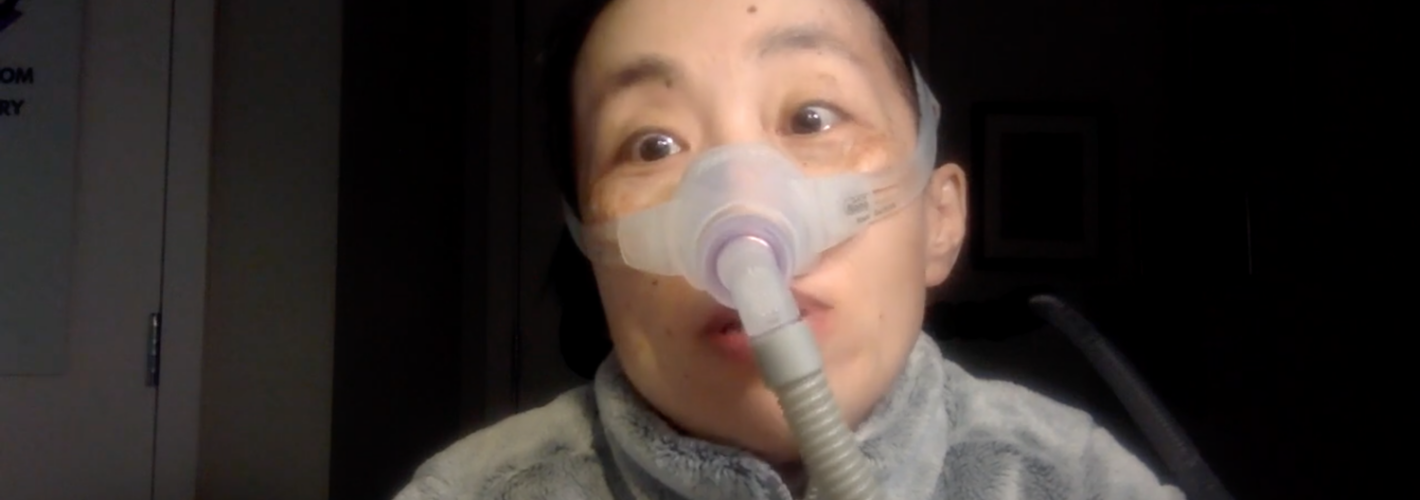 Image description: a picture of Alice Wong, Asian American woman in a wheelchair with short black hair. She is wearing a fleece gray jacket and wearing a mask over her nose connected to a ventilator. The background is a darkened room