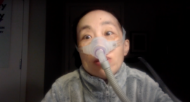 Image description: a picture of Alice Wong, Asian American woman in a wheelchair with short black hair. She is wearing a fleece gray jacket and wearing a mask over her nose connected to a ventilator. The background is a darkened room
