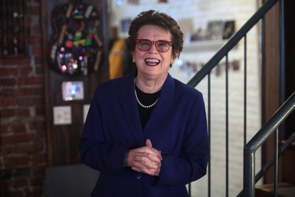 Image of Billie Jean King, an older white woman, smiling and laughing. She is wearing a blue suit jacket and red glasses.