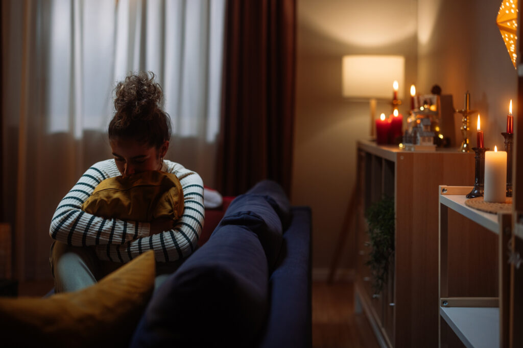 A grieving woman sits alone on the couch, hugging a pillow, in a dark room lighted by candles.