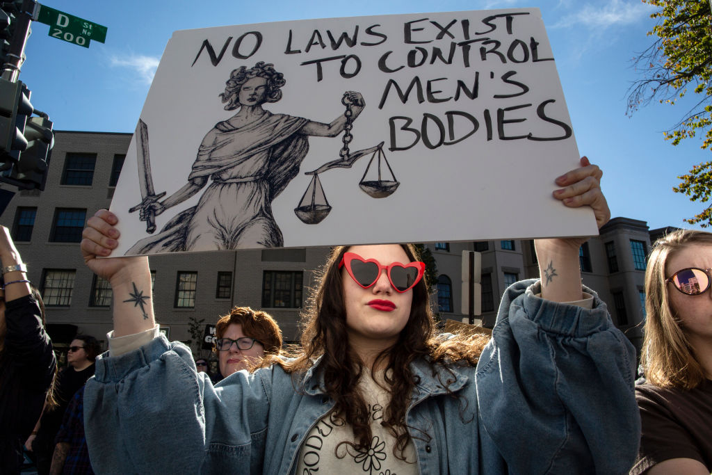 activist holding a protest sign with "No Laws Exist to Control Men's Bodies" written on it