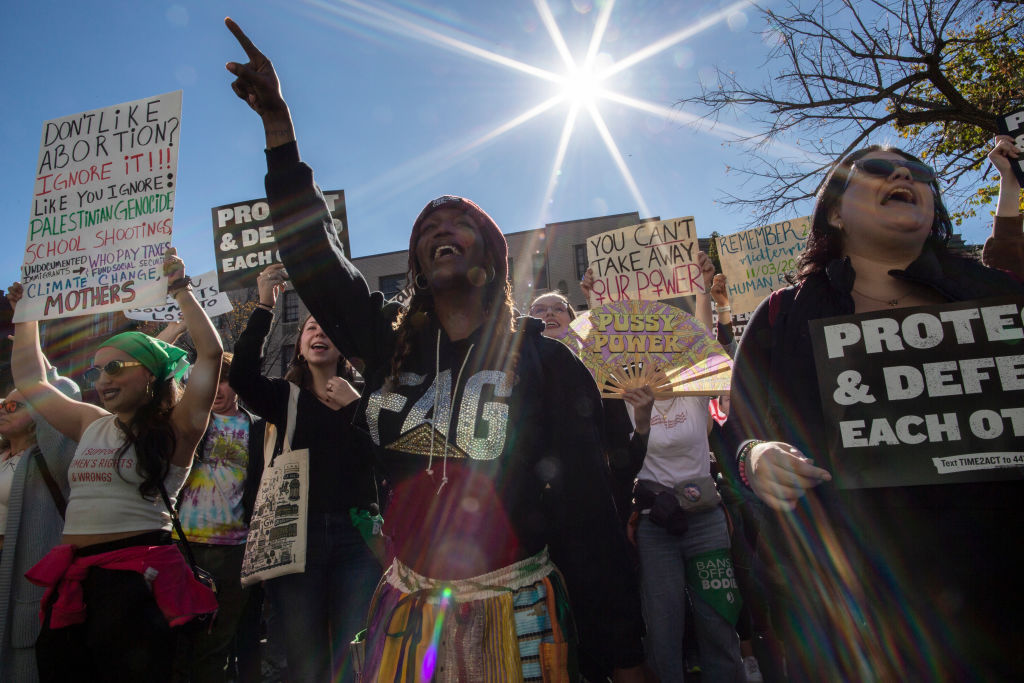 Abortion rights activists stand under a clear blue sky, holding protest signs and singing with a live song.