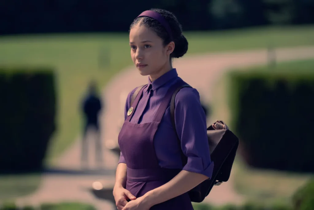 Actress Chase Infiniti, a character in Margaret Atwoods novel The Testaments, stands looking off camera in a purple dress, carrying a backpack. 