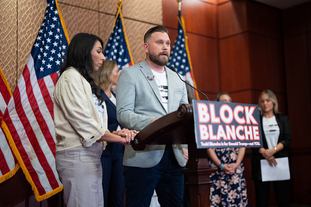 A man in a white t-shirt and a gray blazer stands at a wooden podium displaying a sign that says "BLOCK BLANCHE." Behind him are four women and a row of American flags. 