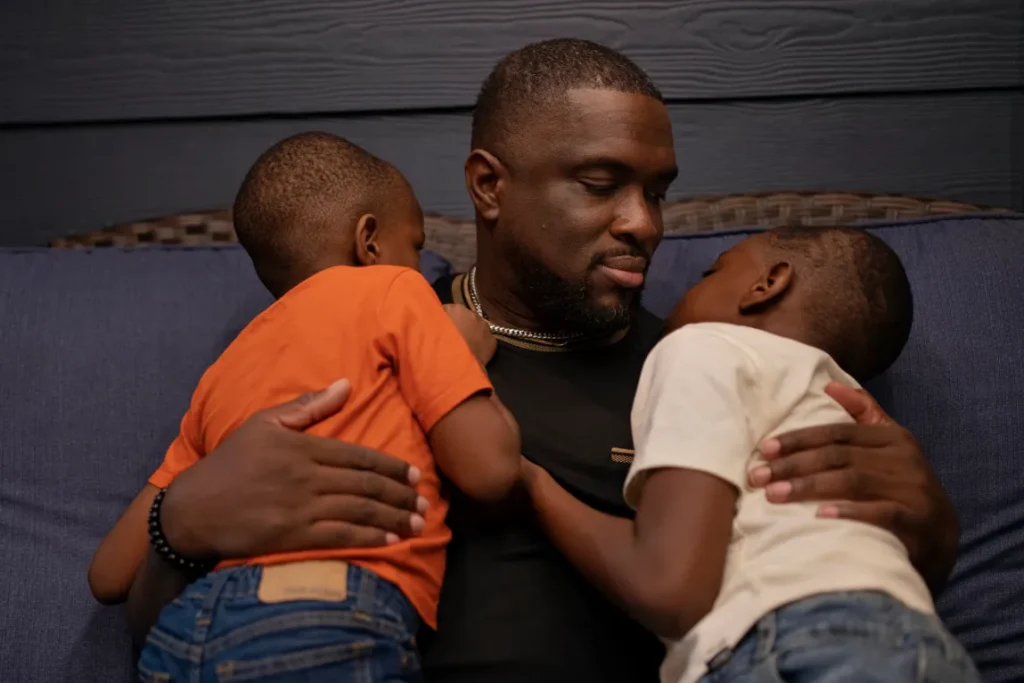 A Black man sits on a blue couch holding his two sons, one wearing orange and one wearing white. 