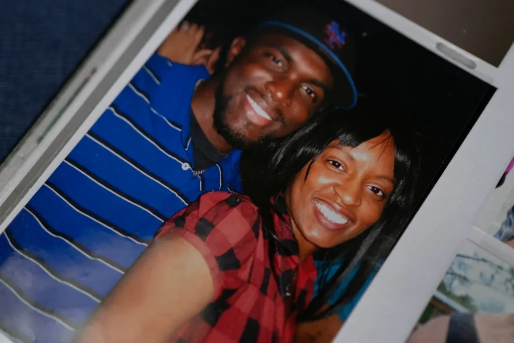 A Black man in a blue striped shirt and a black baseball cap stands next to a Black woman with dark hair and a red and black plaid top. They are smiling. 