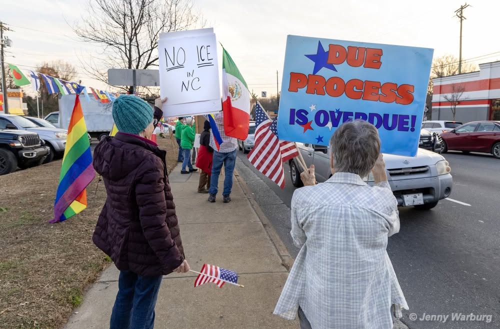 A crowd of protesters line the street holding pride flags and American flags. Their signs say, "NO ICE IN NC" and "DUE PROCESS IS OVERDUE!"