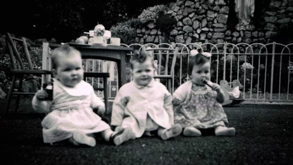 In a black and white photo, three infant children sit in front of a wooden table and a rock wall. 