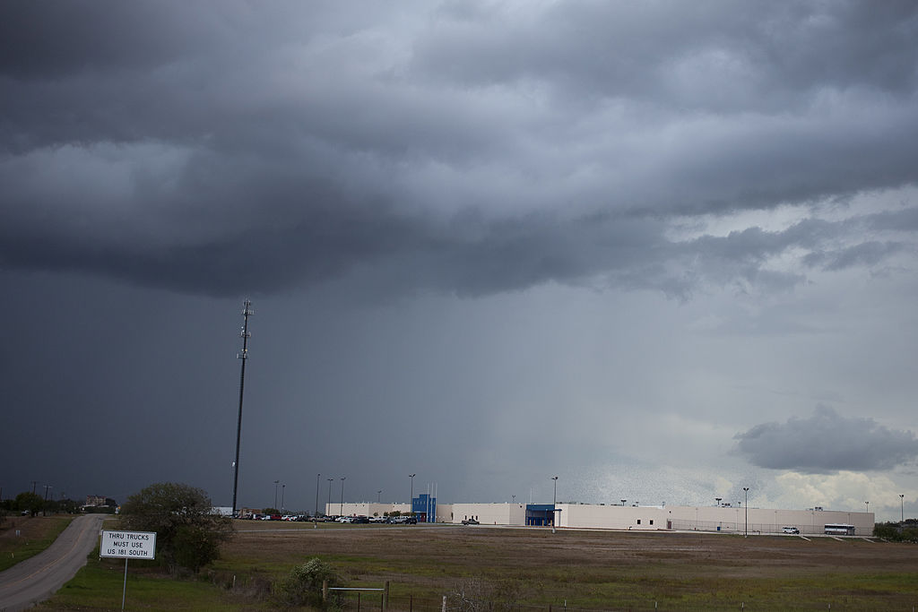 The sky is gray and stormy over a flat white building with a field in front of it: The Karnes County Civil Detention Center. 