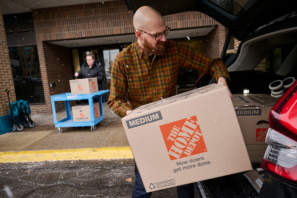 It snows as a bald man in a yellow and orange plaid shirt loads a Home Depot cardboard box into the back of a car in front of a brick building.