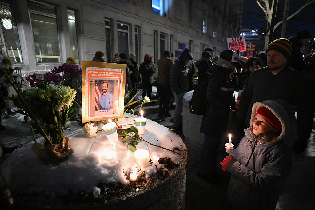 A child red gloves and a winter coat holds a candle as she looks at a vigil with a photo of Alex Pretti.
