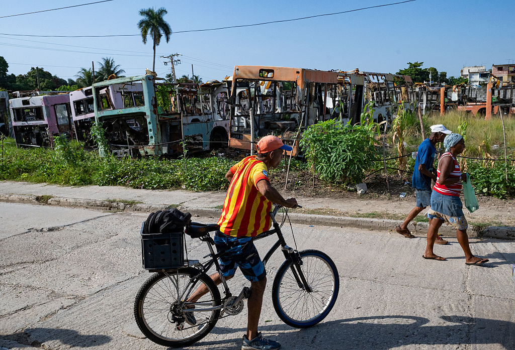 People walk past a man in a striped shirt on a bike in front of bus carcasses in Havana.