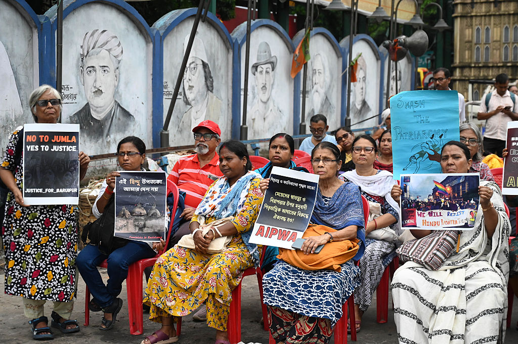 A group of Indian men and women sits on red chairs in front of a wall of murals. They hold signs in protest against the rape and murder of a local girl.