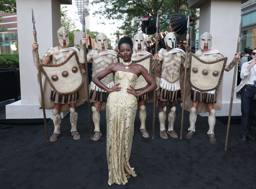 Black actress Lupita Nyong'o is dressed in a gold gown, standing in front of men dressed as Trojan soldiers at the premier of The Odyssey film.