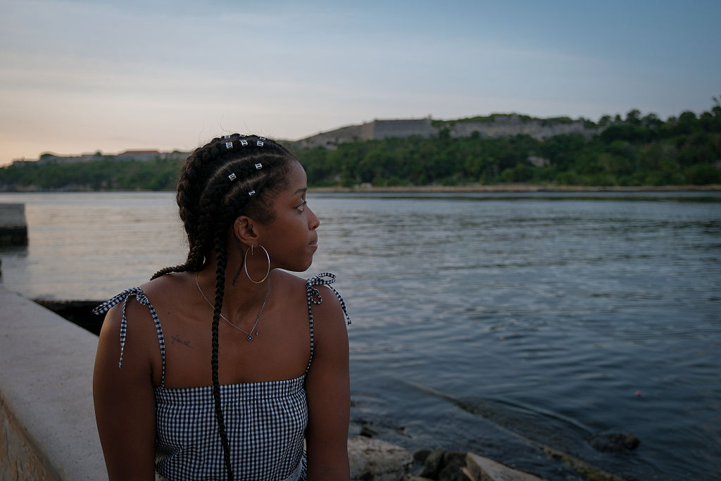A young Cuban woman with braided hair and wearing a checkered top looks out onto a lake.