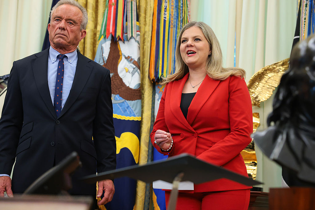 Dr. Heidi Overton, a blond woman in a red pantsuit, stands next to RFK Jr., a greying man in a blue and navy suit, as they speak to a crowd in the Oval Office.