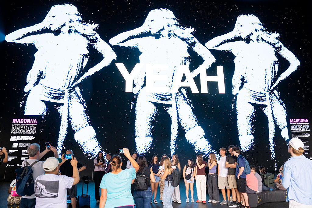 A crowd gathers in the foreground to take pictures of a digital display of a repetitive image of Madonna with the word "YEAH."