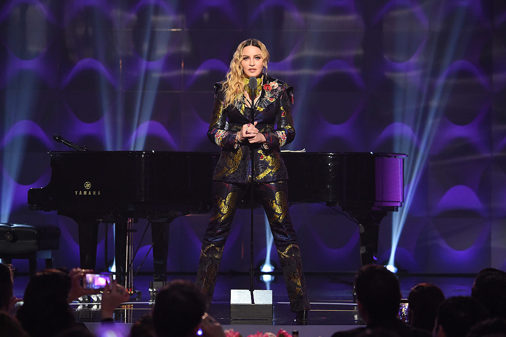Madonna stands in front of a piano with long blond hair and a multi color suit as she sings into a microphone.