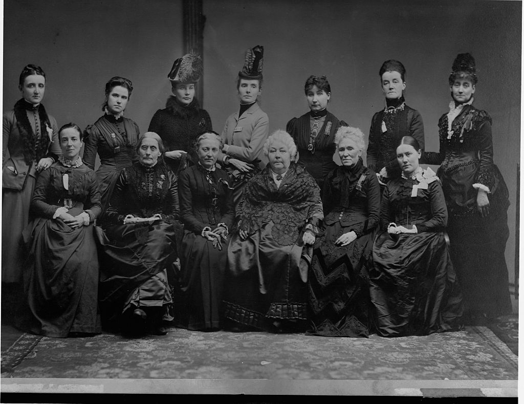 In a black-and-white image, a group of white women sit and stand for a photo. They wear traditional dresses from the 19th century. 