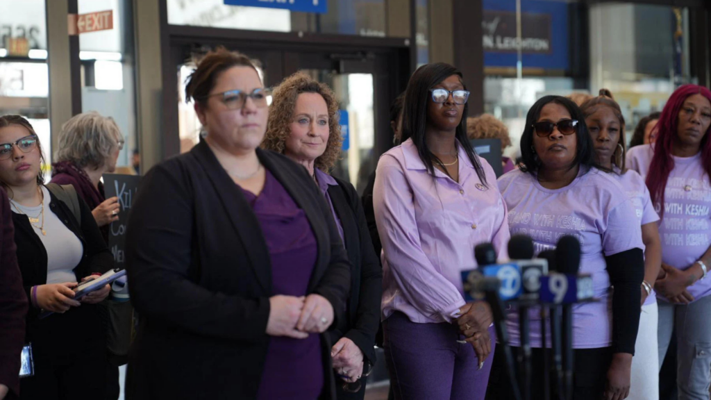 A group of women in different colors of purple stand alongside each other in front of a set of microphones. 