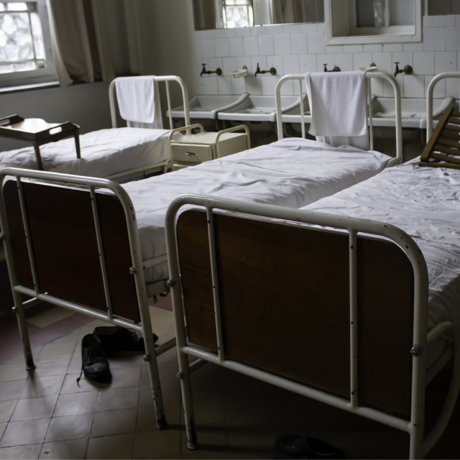 A row of three institutional hospital beds from the 20th century sit in front of a row of sinks. 