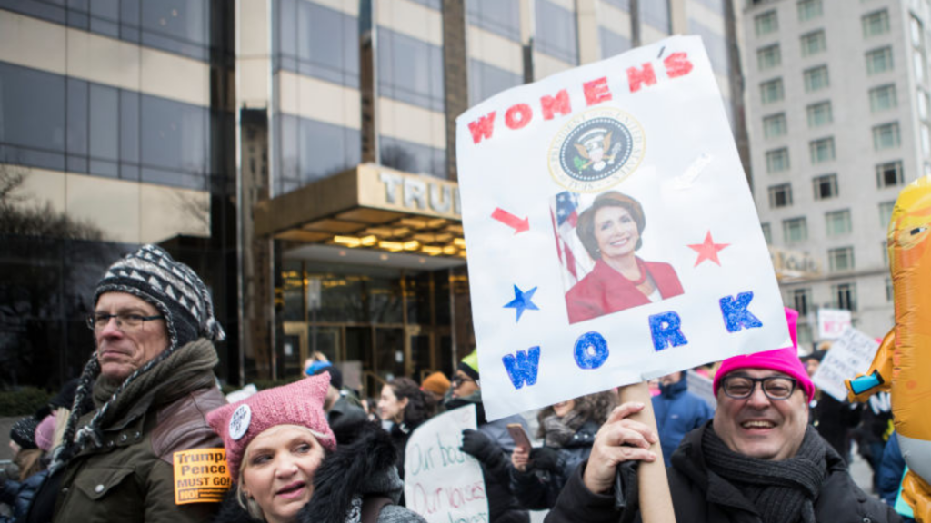 A marcher wearing a pink hat with a sign that says, "Women's Work" with a picture of Nancy Pelosi, Speaker of the United States House of Representatives and holding a balloon of Donald Trump in a diaper.