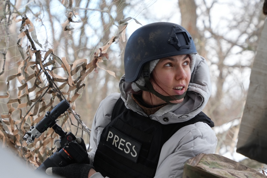 A female front-line journalist wearing a "press" vest and helmet looks back over her shoulder.