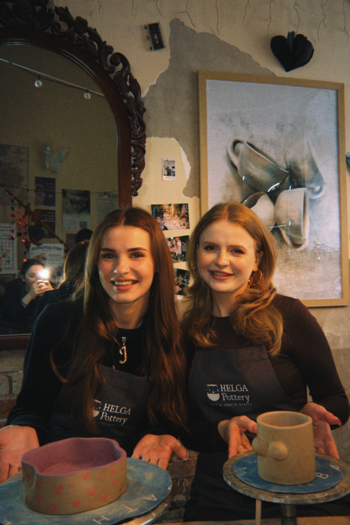 Two women wear their hair down with black HELGA Pottery aprons as they smile at the camera and hold pottery they have made.