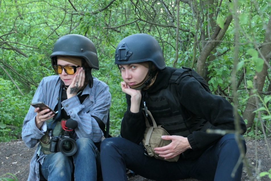 Two female front-line journalists sit on the ground together wearing vests and helmets.