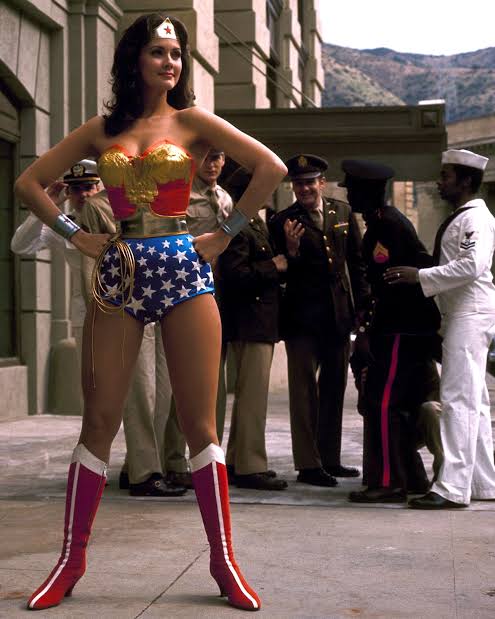Lynda Carter, dressed and acting as Wonder Woman, stands in her charcteristic pose and is oogled by men in the background.