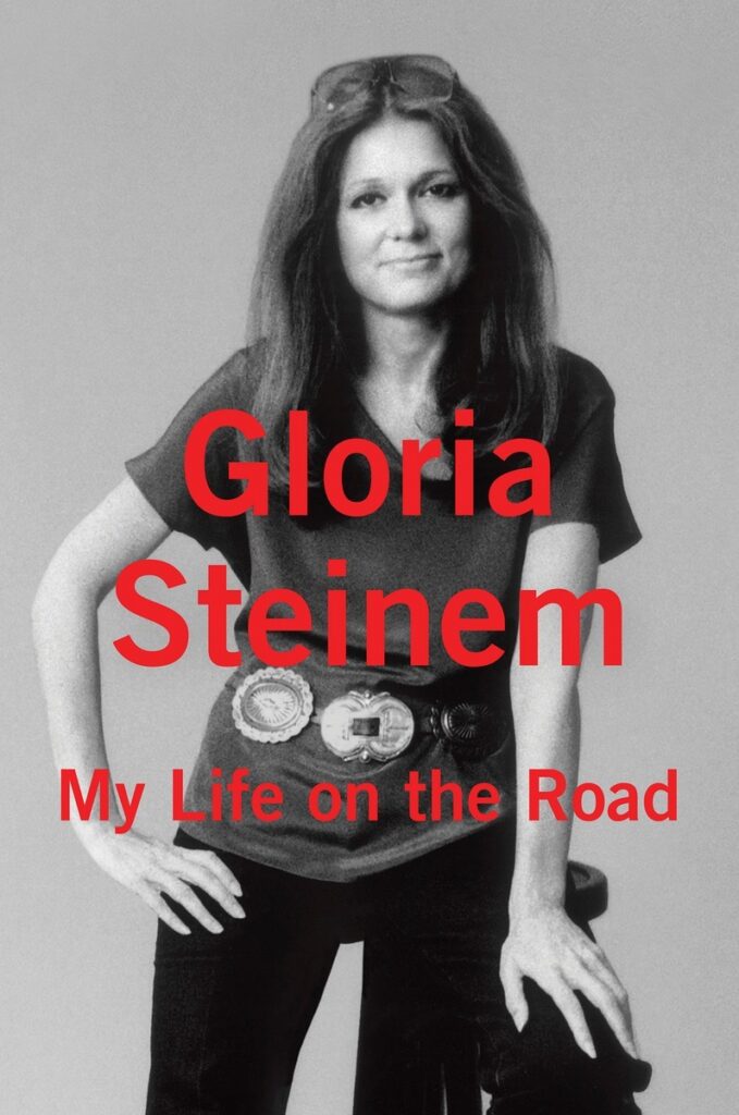 Young Gloria Steinem wears her hair long in this black and white photo of her smirking at the camera. Red lettering on the image says "Gloria Steinem My Life on the Road."