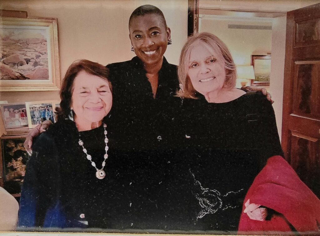 Dolores Huerta, Michele Goodwin and Gloria Steinem stand together smiling in a living room.