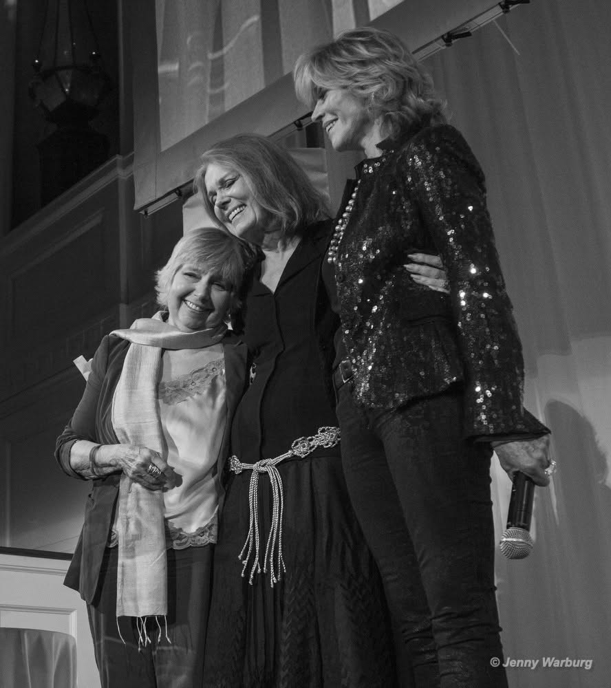 In this black and white photo, Gloria Steinem smiles while side-hugging two women, Robin Morgan and Jane Fonda.