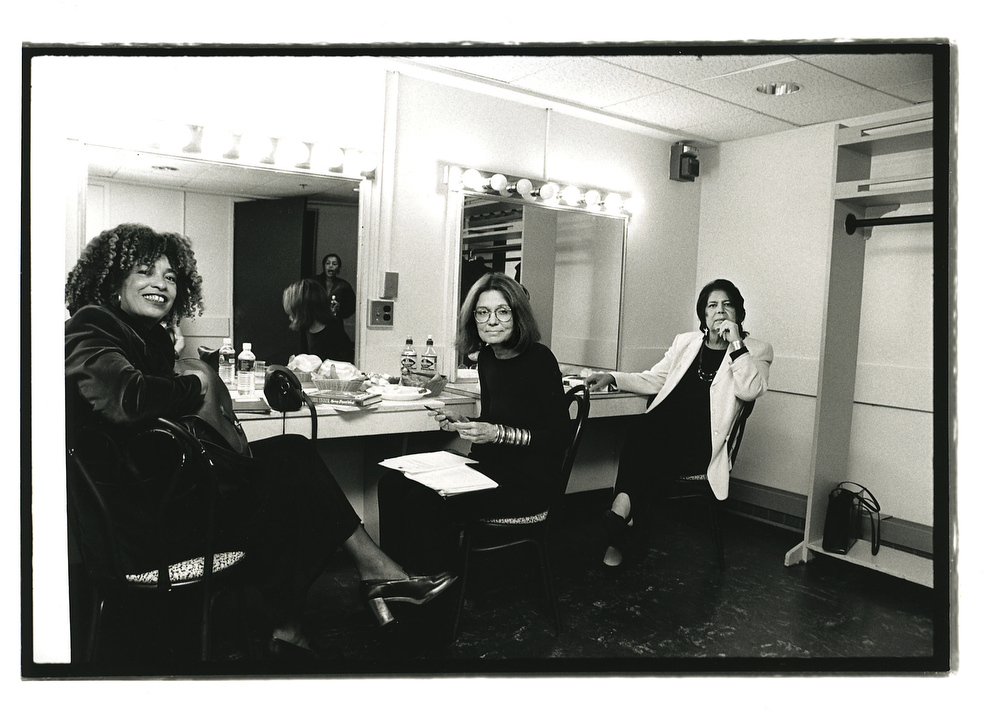 A black and white photograph is taken of a young Gloria Steinem, flanked by Angela Davis and Wilma Mankiller as they sit together at lit mirrors. 