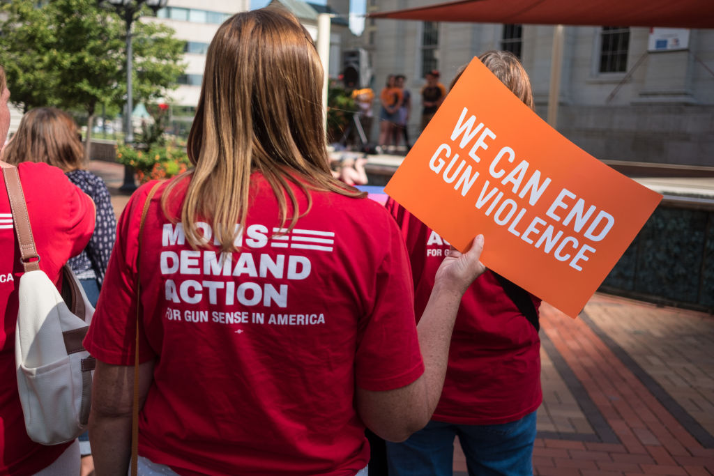 A woman wearing a red t-shirt holds an orange placard that says "WE CAN END GUN VIOLENCE."