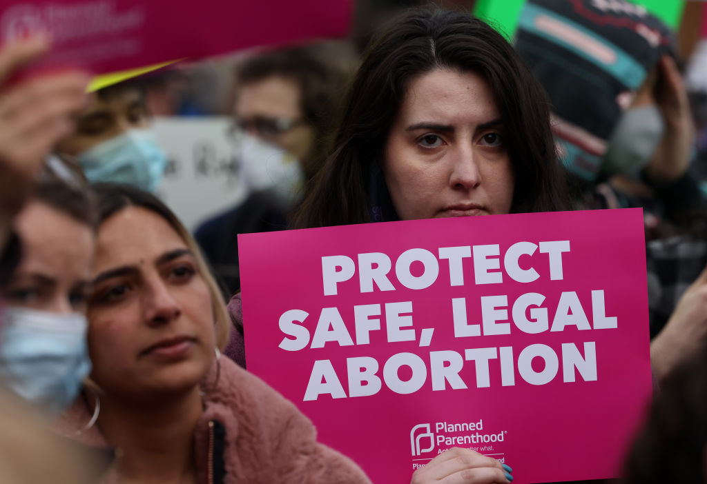 A protester with long brown hair holds a pink sign that says "PROTECT SAFE, LEGAL ABORTION."