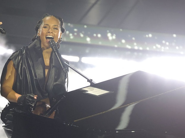 Alicia Keys sits on a backlit stage wearing black and singing into a microphone attached to a piano.