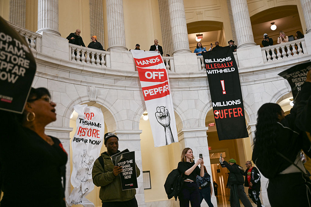 In the Cannon House Office Building in Washington, D.C., people protest and hang signs off the balconies that say "Hands off PEPFAR" and "Billions Illegally Withheld Millions Suffer." 