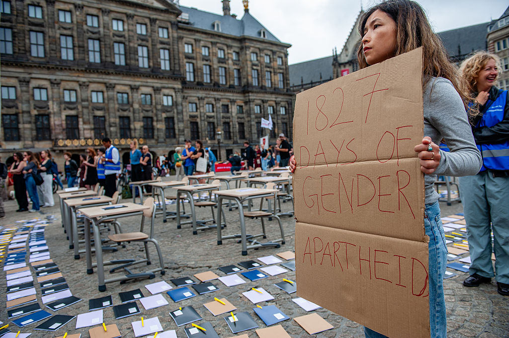 A woman at a demonstration with desks and notebooks sitting in a courtyard holds a cardboard sign that says "1827 DAYS OF GENDER APARTHEID."