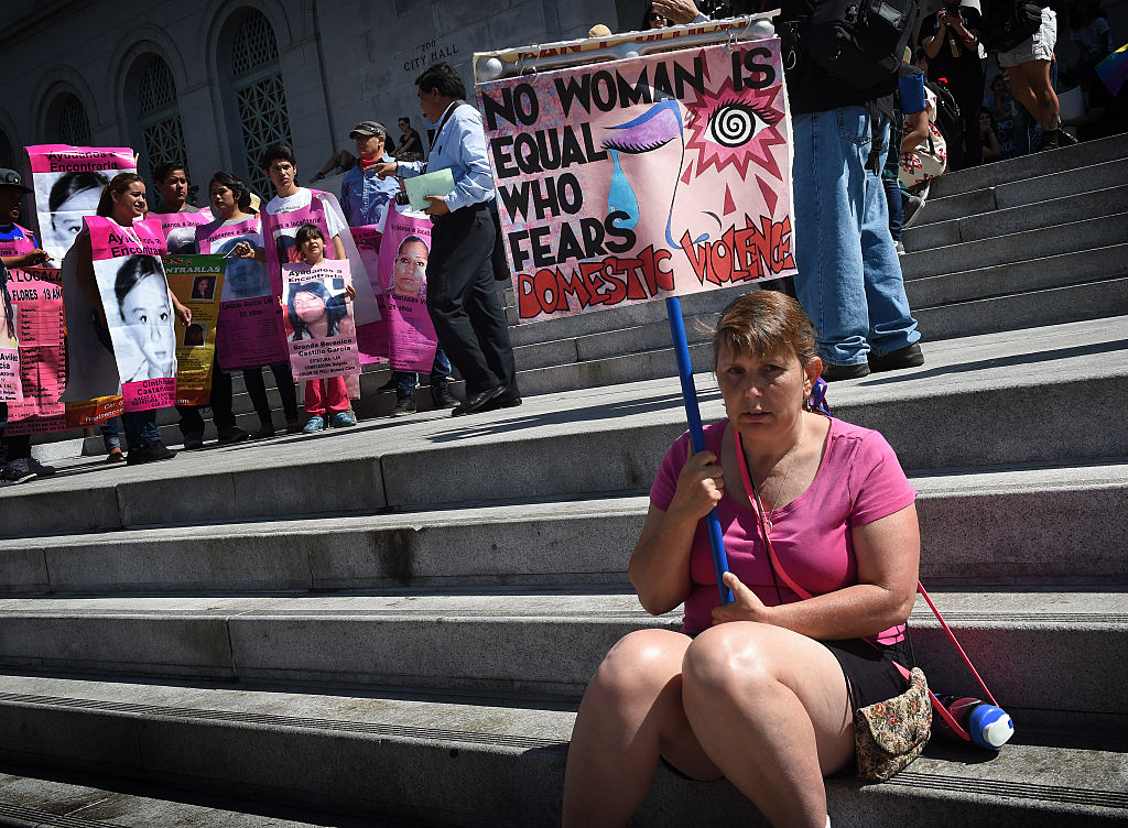 A woman wearing pink sits on steps in Los Angeles and holds a sign that says "NO WOMAN IS EQUAL WHO FEARS DOMESTIC VIOLENCE."