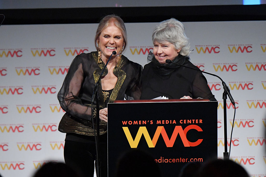 Gloria Steinem, wearing a black and gold top, stands at a podium with Robin Morgan, wearing a black turtleneck top, at the Women's Media Center.