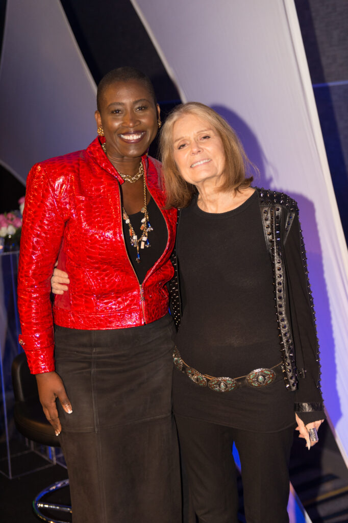 Michele Goodwin, a Black woman with short hair and a red leather jacket, smiles as she stands with Gloria Steinem, wearing a black studded jacket.