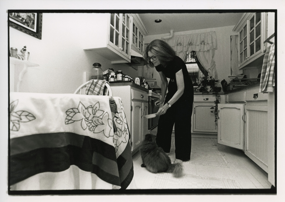 In this black and white photo, Gloria Steinem bends over in her kitchen to feed her fluffy cat.