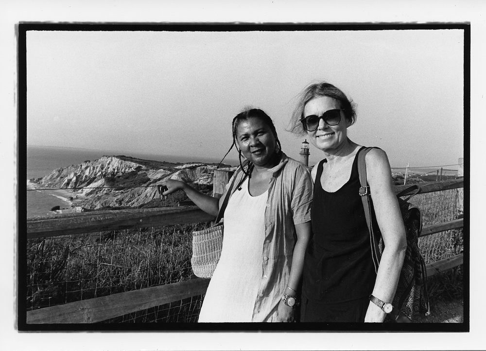 In this black and white photo, Gloria Steinem and bell hooks stand together smiling in front of a coastal view.