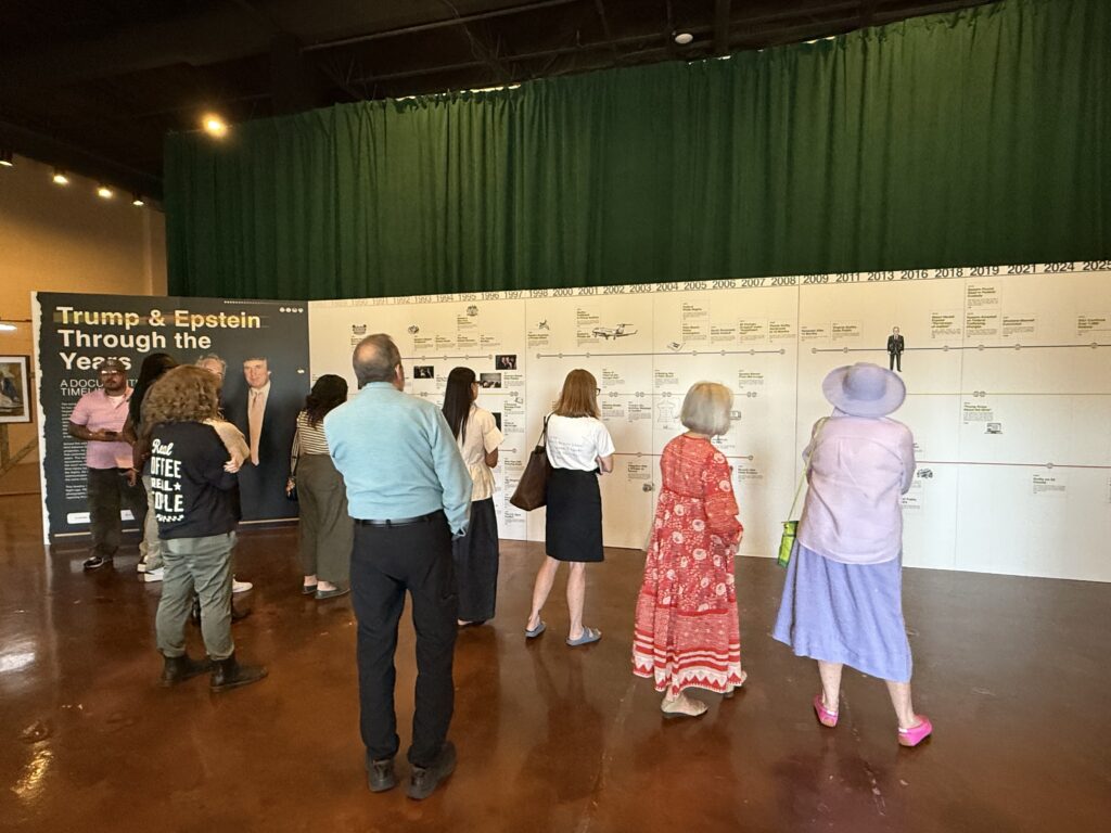 Men and women stand in front of a wall displaying a timeline at the Donald J. Trump and Jeffrey Epstein Memorial Reading Room exhibit.