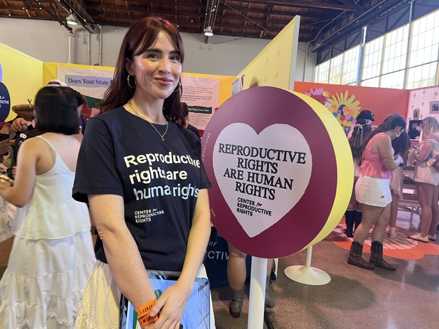 A woman with long hair wears a black t-shirt that says "Reproductive rights are human rights" and stands next to a circular pink sign that says "REPRODUCTIVE RIGHTS ARE HUMAN RIGHTS" in a heart.