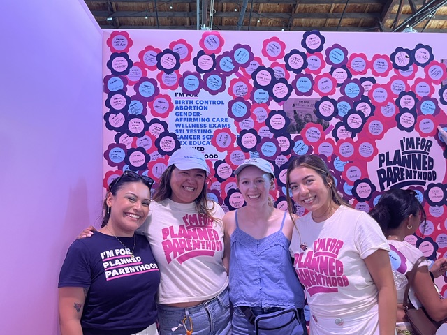A group of four women wearing Planned Parenthood shirts stand in front of a wall of handwritten notes.