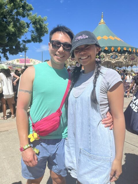 Dale Yu, with short hair and a turquoise tank top on, and Abby Louie, with a blue jean dress and black baseball hat on, stand together smiling.