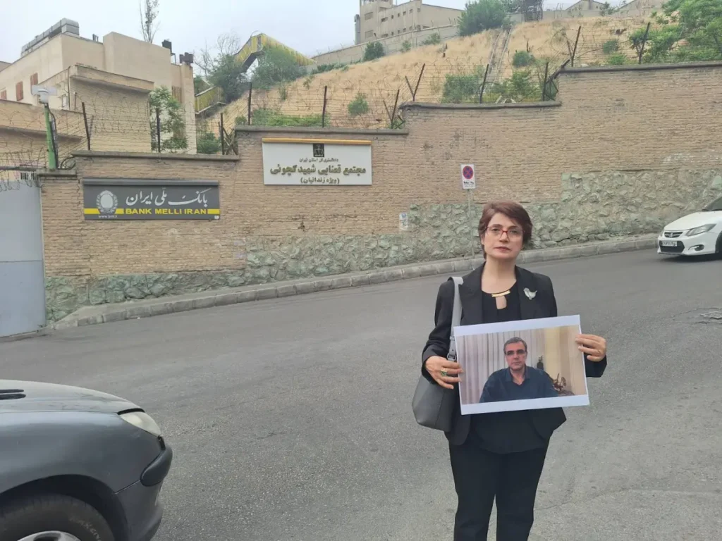 Nasrin Sotoudeh, a woman with pixie-cut hair, glasses and wearing a black suit, stands in front of a brick wall holding a photograph of Reza Khandan. 