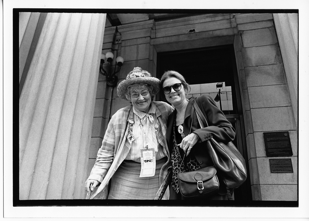 Gloria Steinem wears a blazer and sunglasses as she smiles with Bella Abzug, a woman with short hair wearing a floral hat.